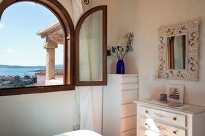 Ein Schlafzimmer mit großem Fenster, Kommode, Spiegel und einer Vase mit Trockenpflanzen.