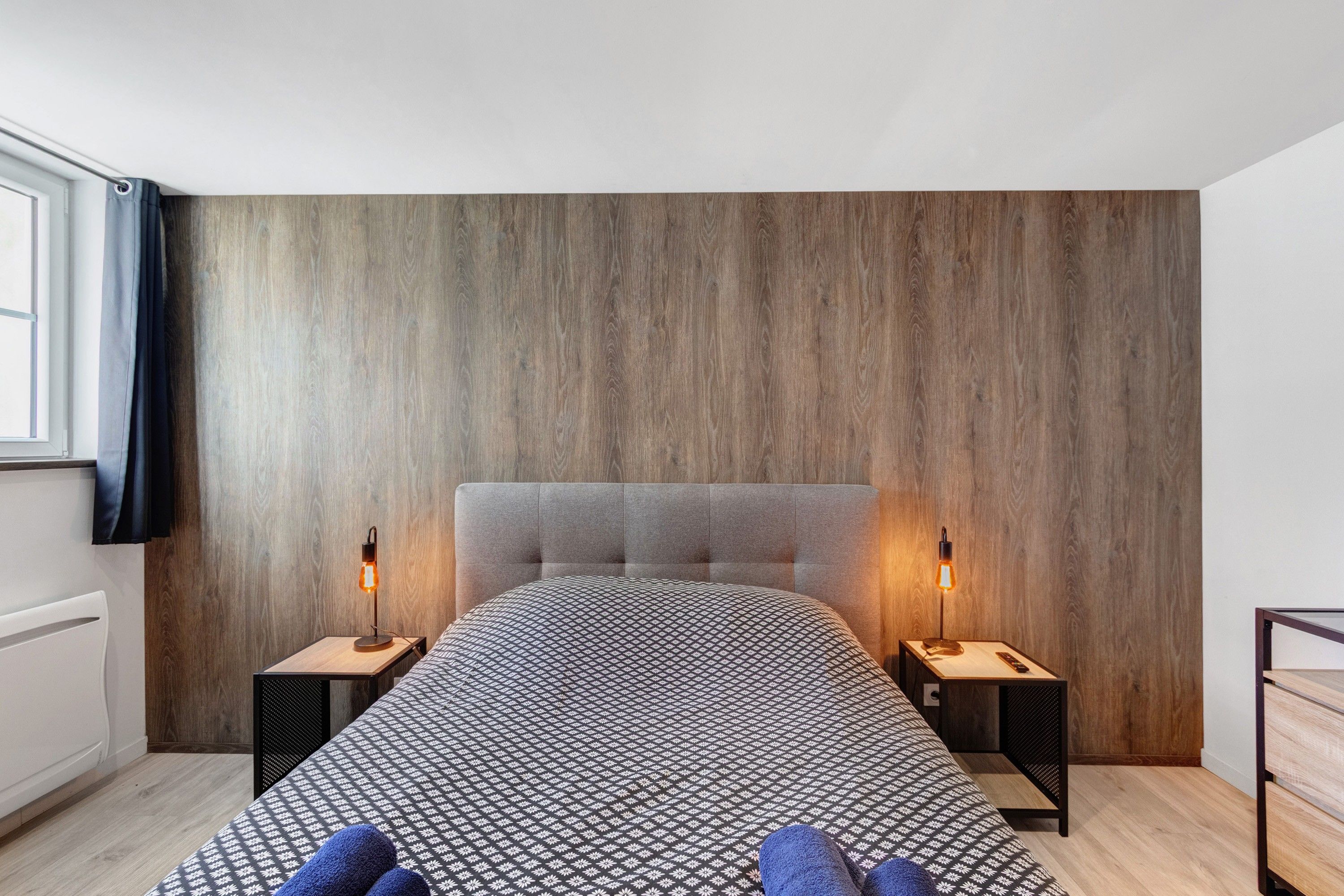 A modern bedroom with a wooden headboard, a patterned bedspread, and two bedside lamps.