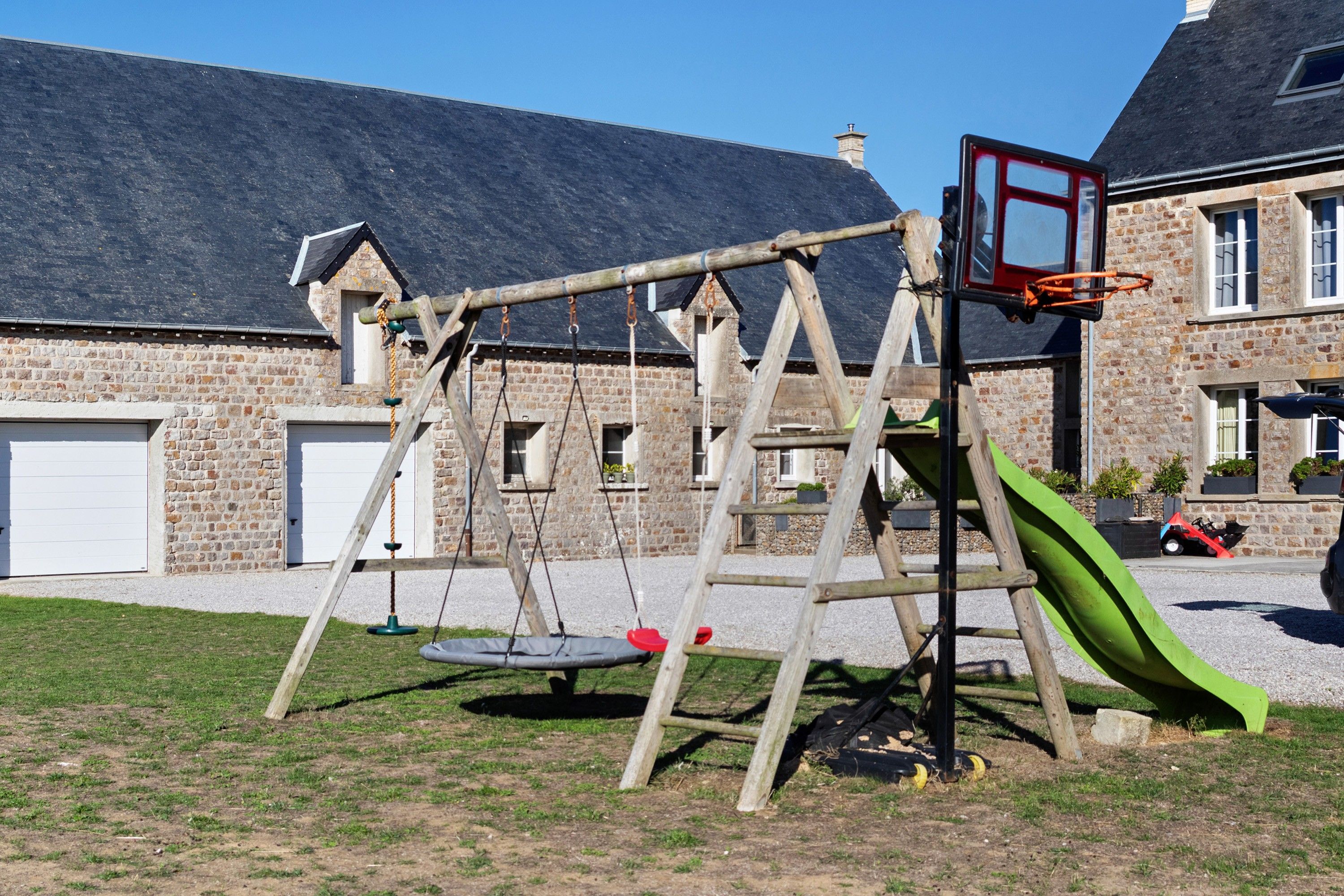 Een houten schommel met een glijbaan, een basketbalveld en een garage op de achtergrond.