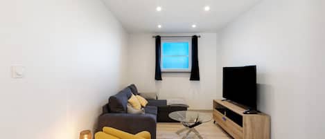 Una sala de estar moderna con un sofá, un sillón amarillo, una mesa de centro de vidrio, un televisor sobre un mueble de madera y una ventana con cortinas.