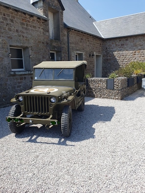 A vintage military jeep parked in front of a stone building with a gravel driveway.