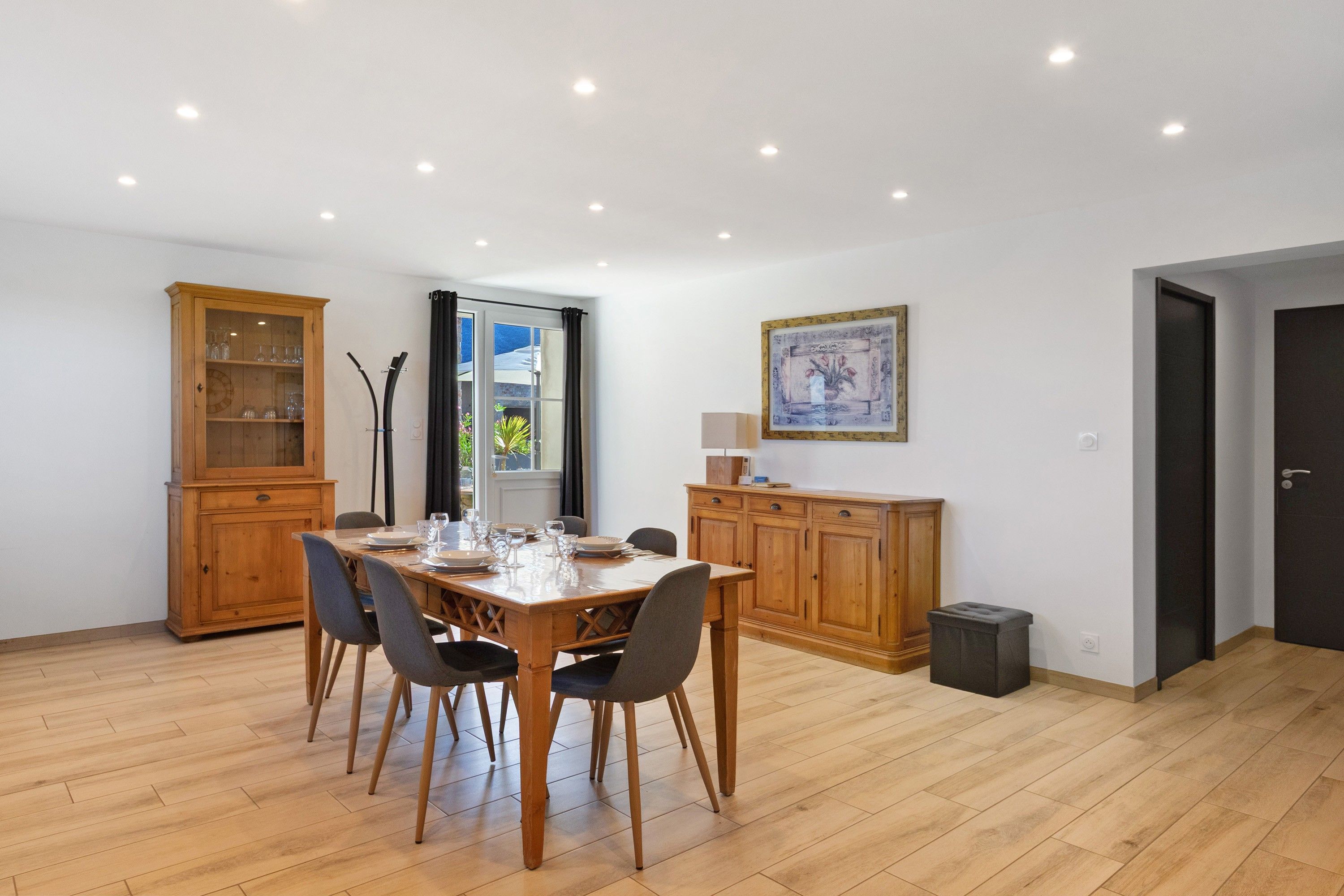 Un coin repas avec une table en bois dressée pour quatre personnes, bordée de meubles en bois, une fenêtre avec des rideaux et un tableau encadré au mur.
