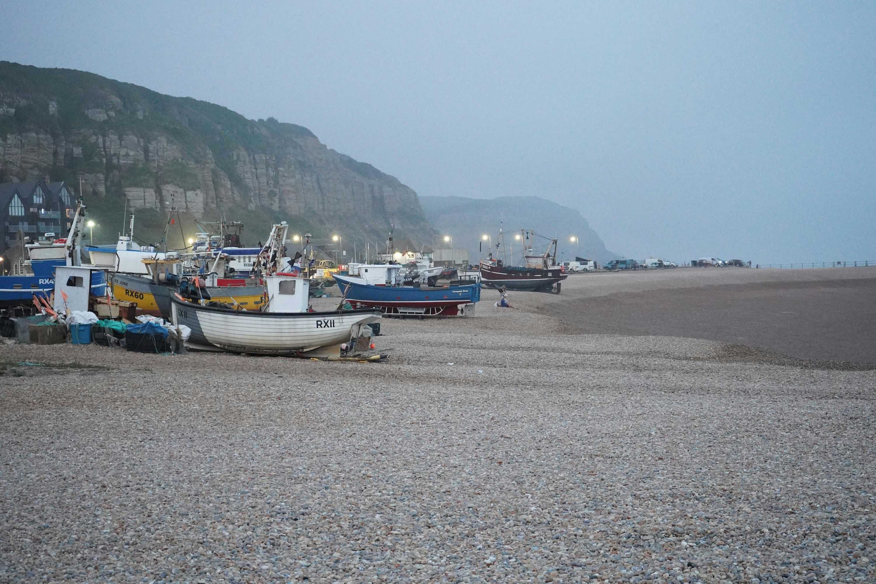 A pebbled beach with boats docked, a cliff in the background, and a clear sky.