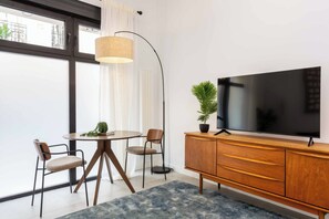 A modern living room with a flat-screen TV, wooden cabinet, two chairs, a round table, and a floor lamp. - A spacious furnished studio in the heart of Tetuán with its own private patio, for slow coffee in the morning or a workday in the sun. (MADRID)