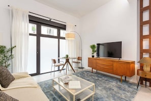 A modern living room with a sofa, a coffee table with a book, a TV on a wooden cabinet, and a dining area with chairs and a table. - A spacious furnished studio in the heart of Tetuán with its own private patio, for slow coffee in the morning or a workday in the sun. (MADRID)