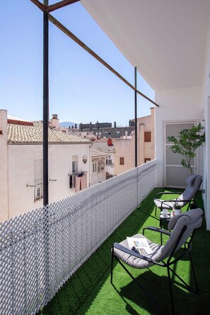 Ein Balkon mit Kunstgrasanlage, zwei Stühlen, einem weißen Zaun und Blick auf Gebäude.