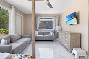 Ein Zimmer mit einer Couch, einem an der Wand befestigten Fernseher, einem Holzschrank und einem Ventilator.