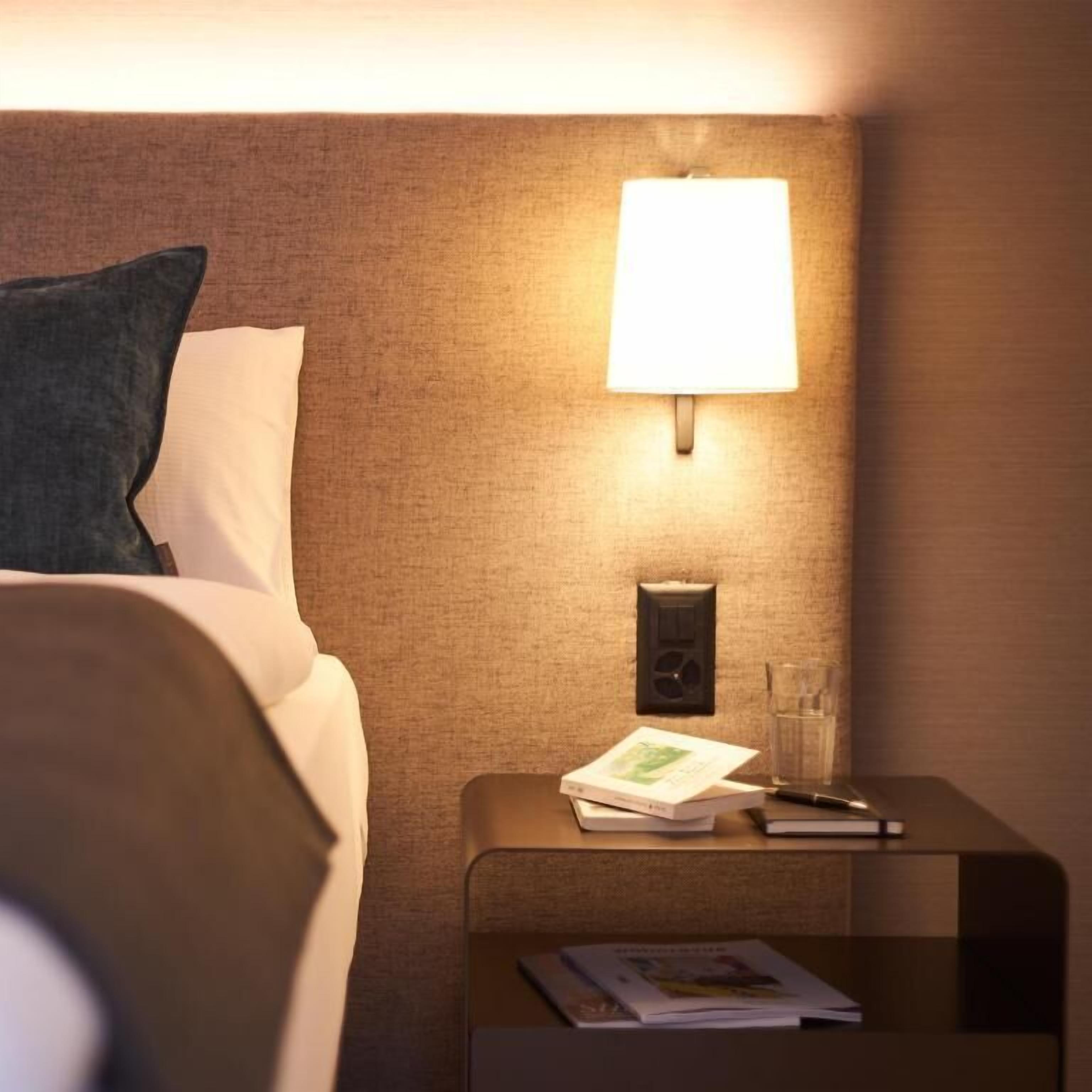 Ein ordentlich gemachtes Bett mit Kissen, ein Nachttisch mit Lampe, einem Buch und einem Glas Wasser.