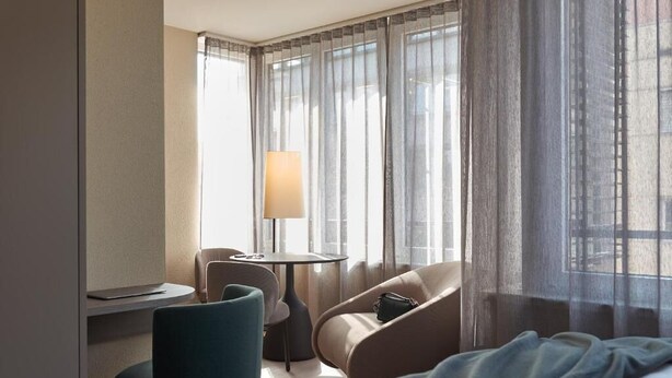 Habitación de hotel con cama, escritorio, silla, lámpara y vistas a través de cortinas translúcidas.