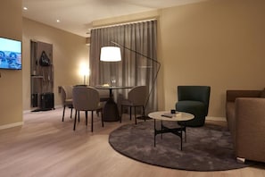 Quarto de hotel moderno com área de refeições, sofá, televisão e uma grande janela com cortinas.