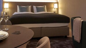 Habitación de hotel con cama, lámparas de noche, una mesa redonda con una silla y una mesita con una copa de vino y un plato.