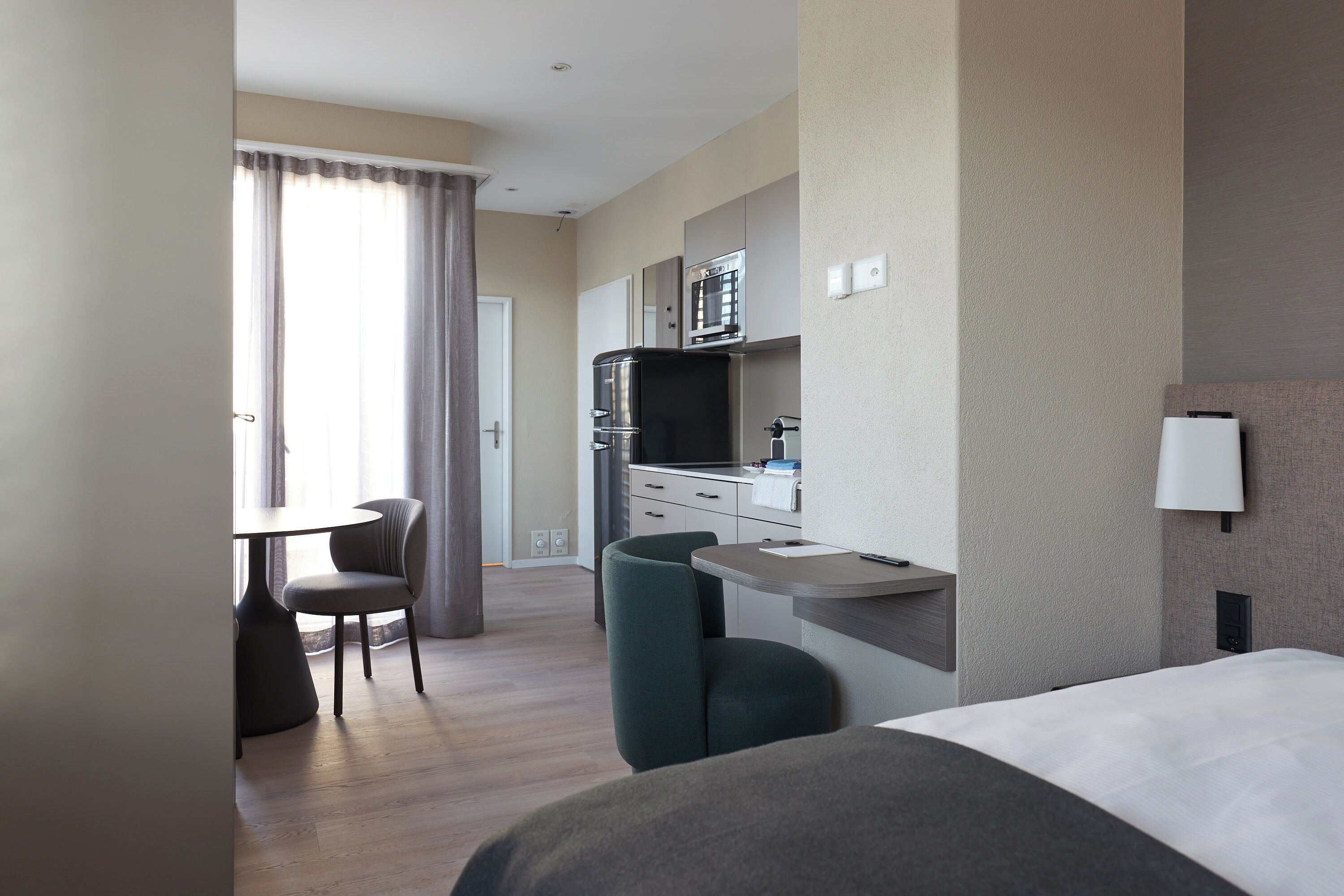 A hotel room with a bed, a desk, a chair, a TV, and a kitchenette.