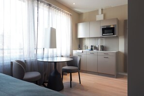 Una habitación de hotel moderna con zona de comedor, cocina con microondas y un ventanal con cortinas translúcidas.