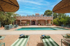 Un área de piscina con sillones de descanso y sombrillas, un edificio con arcos, y cielo despejado.