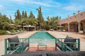 Una piscina con sillas de descanso y sombrillas, rodeada de árboles y un edificio con arcos.