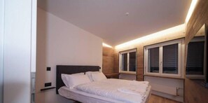 Een slaapkamer met een bed, een houten hoofdbord, twee ramen met zonwering en een lamp boven het bed.