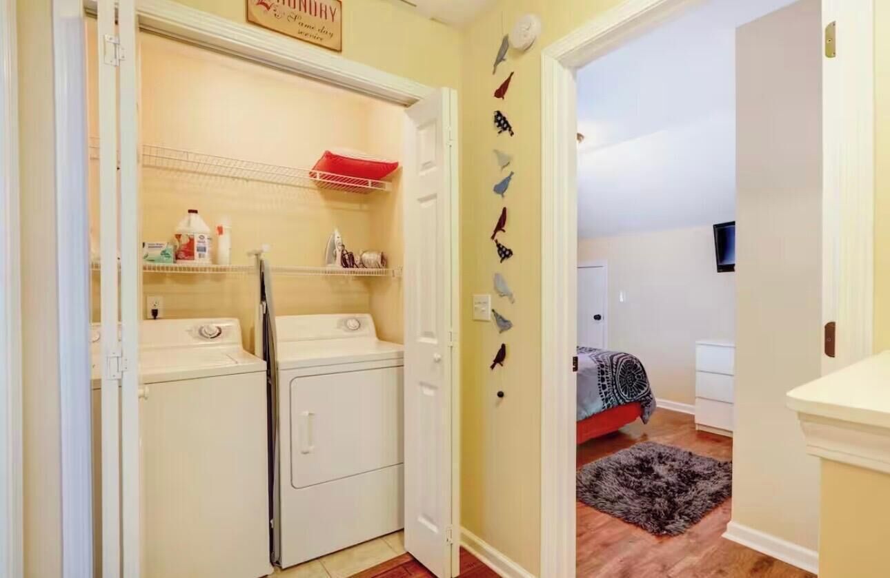 A laundry room with a washer and dryer, a shelf with cleaning supplies, and a door leading to a bedroom with a bed and a rug.