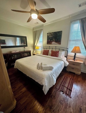 Una cama bien hecha con toallas, un ventilador de techo, un cuadro, una lámpara y una ventana con cortinas.