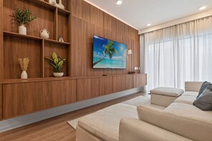 Una sala de estar moderna con una estantería de madera, un televisor de pantalla plana, un sofá beige y un amplio ventanal con cortinas traslúcidas.