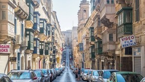 Una strada fiancheggiata da auto parcheggiate e edifici con insegne per 'Video Film Rentals' e 'Argain Books'.