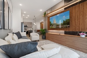 A modern living room with a large flat-screen TV mounted on a wooden wall, a sofa with cushions, a coffee table, and a potted plant. - Fun Family Townhome with Pool and Game Room! (Orlando)