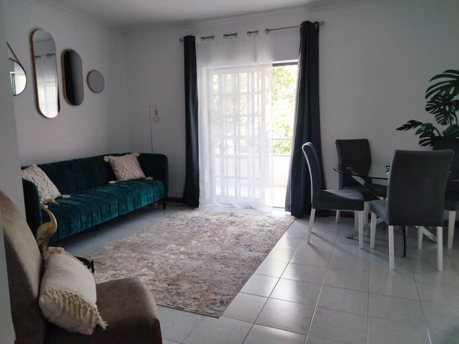 Ein Wohnzimmer mit Sofa, einem Esstisch mit Stühlen, einem großen Fenster mit Vorhängen und einer Zimmerpflanze.
