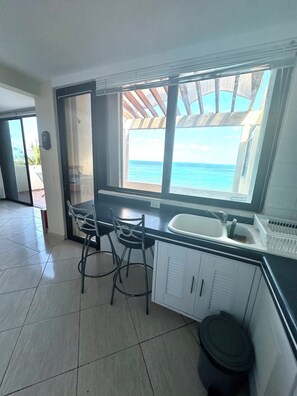 A kitchen with a view of the ocean, a sink, two bar stools, and a trash bin. - Beachfront apartment (Cancún)