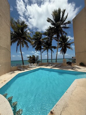 A pool with clear blue water, surrounded by lounge chairs and palm trees, with a view of the ocean. - Beachfront apartment (Cancún)