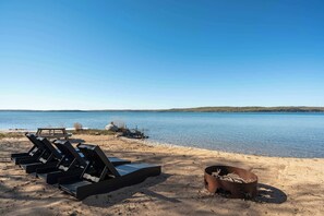A beach with lounge chairs and a fire pit. - Obsidian Shores Waterfront House on Crystal Lake with Private Beach Access (Beulah)