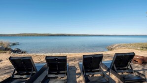 Ein Strand mit vier Liegestühlen, die auf einen ruhigen See ausgerichtet sind, einer Anlegestelle und einer felsigen Uferlinie.