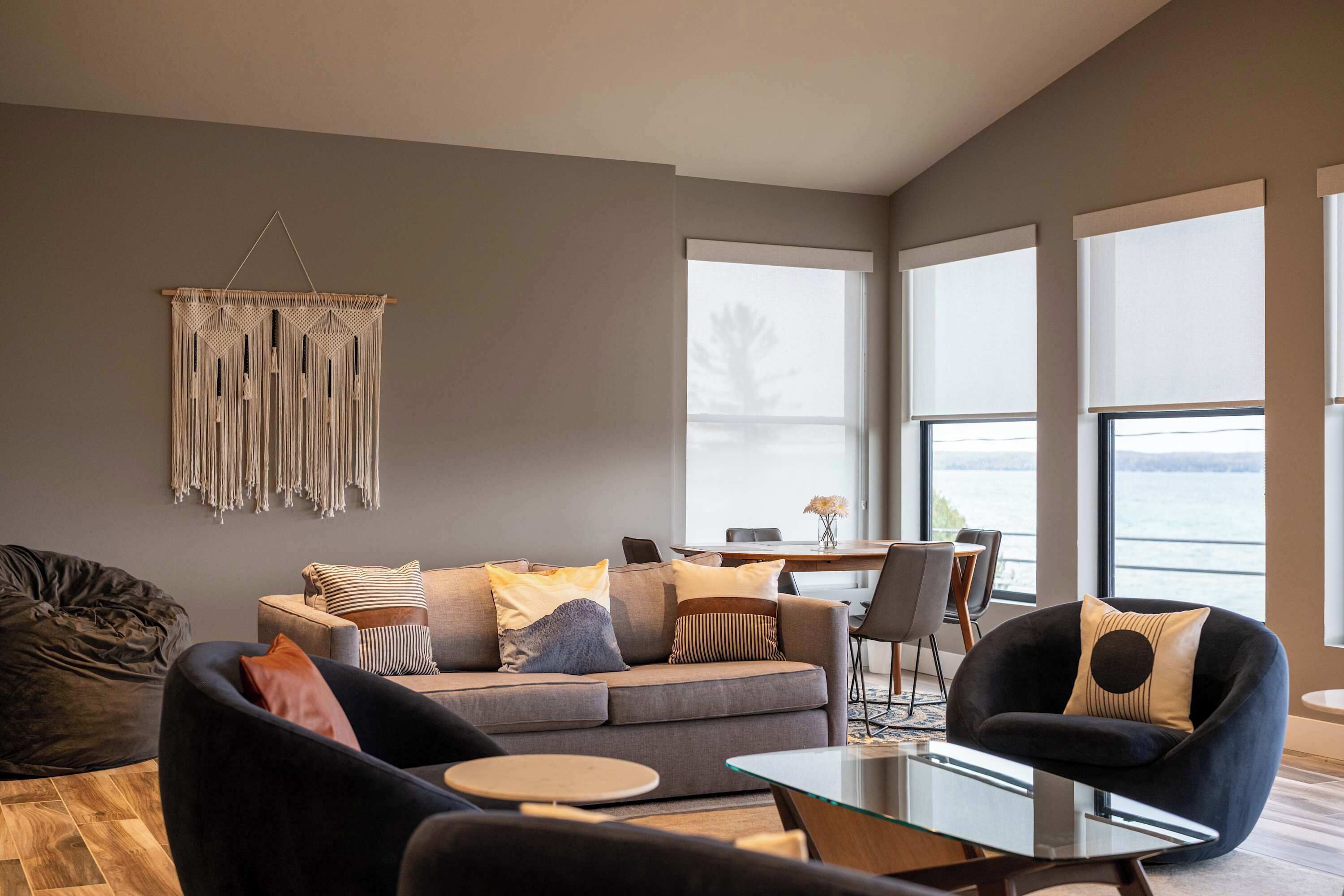 A modern living room with a sofa, armchairs, a glass coffee table, and a macrame wall hanging.