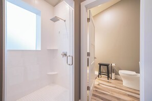 A modern bathroom with a shower, toilet, and a small table with a roll of toilet paper. - Obsidian Shores Waterfront House on Crystal Lake with Private Beach Access (Beulah)