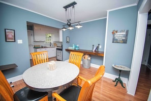 A dining area with a round table, wooden chairs, a kitchen in the background, and a small side table with a lamp.