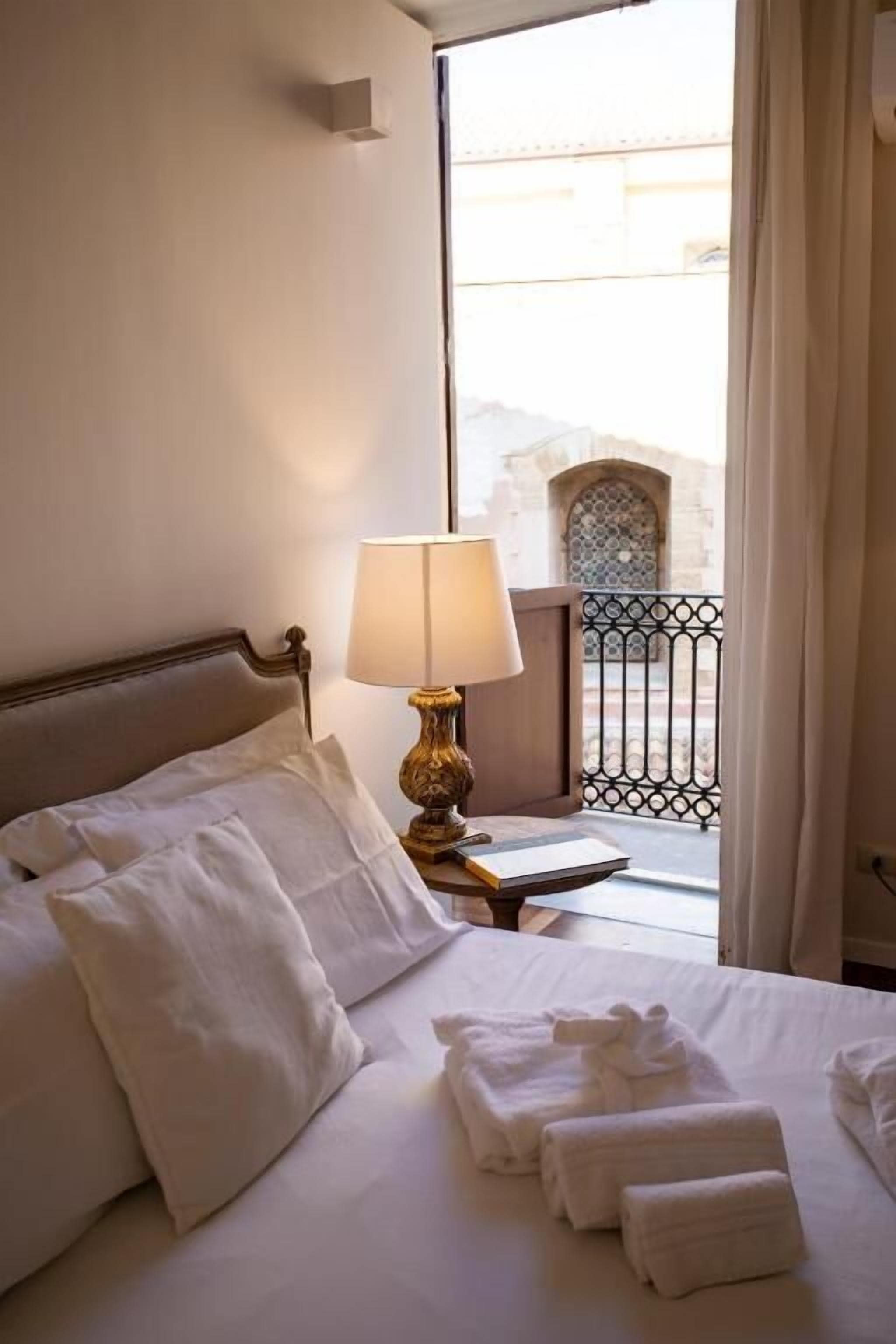 A hotel room with a bed, pillows, a lamp, a window with a view of a building, and a balcony with a railing.