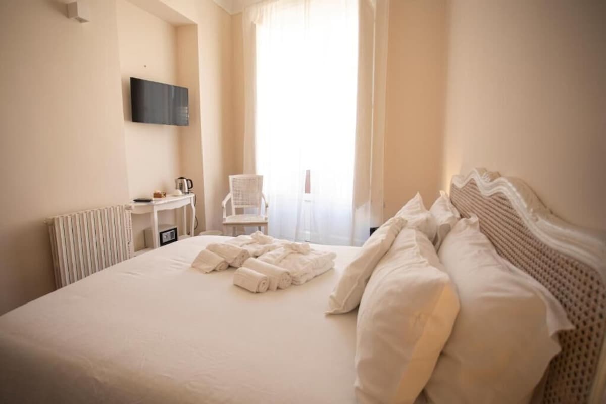 A hotel room with a bed, a chair, a table with a kettle, and a TV mounted on the wall.