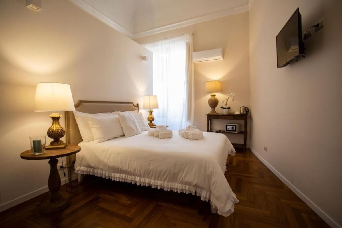 A hotel room with a bed, two bedside tables with lamps, a nightstand with a lamp, a TV mounted on the wall, and a window with curtains.