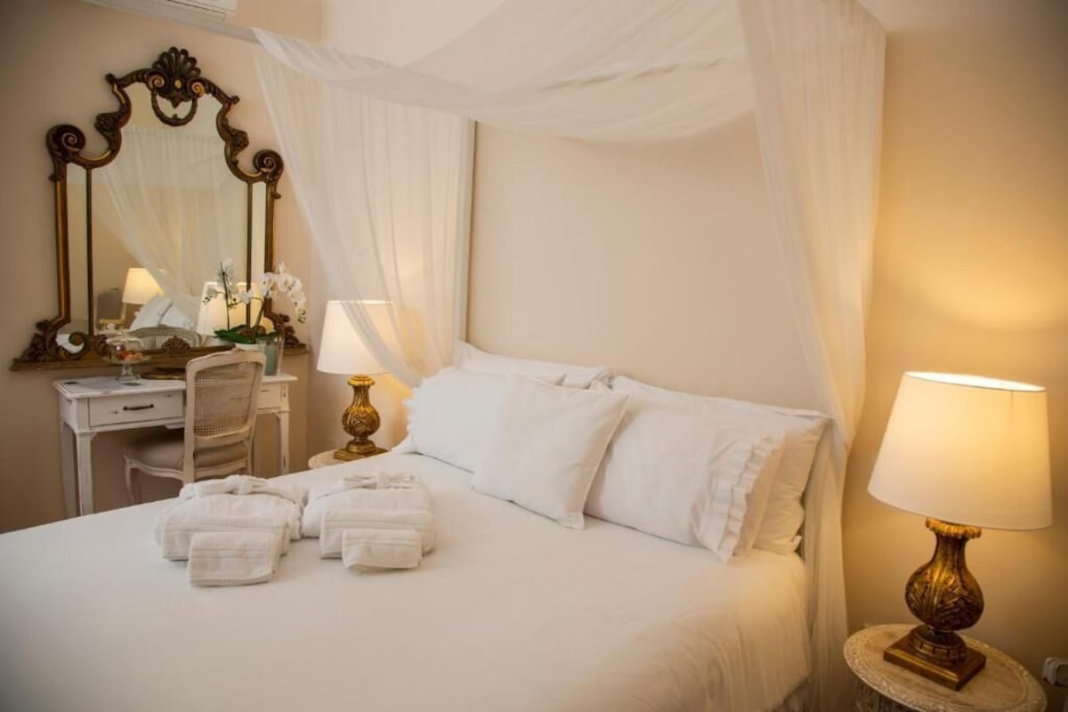 A neatly made bed with white linens, a bedside lamp, and a mirror with a decorative frame.