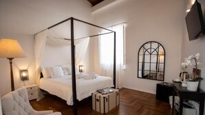 A four-poster bed with white bedding, a bedside lamp, a small bedside table, a chair, a suitcase, a mirror, and a television on the wall. - Dimora Sinibaldi (Palermo)