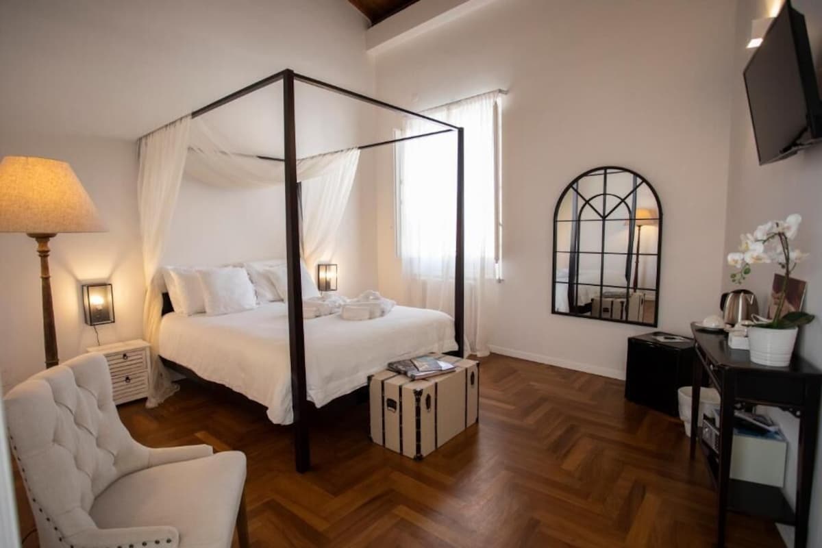 A four-poster bed with white bedding, a bedside lamp, a small bedside table, a chair, a suitcase, a mirror, and a television on the wall.