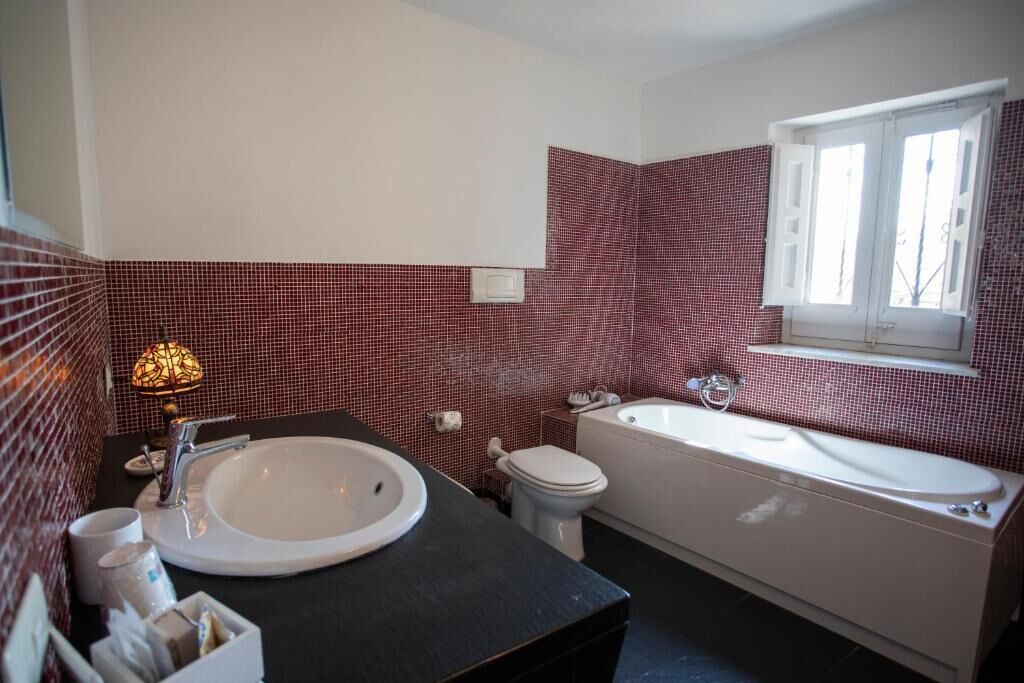 Un bagno con lavandino, WC e vasca da bagno bianchi, pareti rivestite di piastrelle rosse e una finestra.