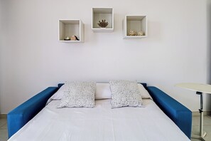 Un llit amb roba de llit blanca i dos almoinos, un sofà blau i tres prestatgeries murals amb objectes decoratius.