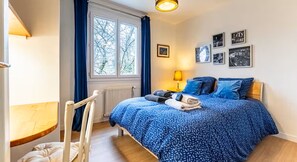 Een slaapkamer met een bed, een raam, een radiator, een stoel, een tafel en wanddecoratie.