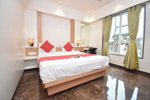 A hotel room with a large bed, a desk, a chair, a mirror, and a window with curtains. - Hotel Royal Oak (Navi Mumbai)