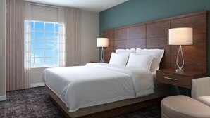A hotel room with a large bed, two bedside tables with lamps, a wooden headboard, a window with curtains, and a carpeted floor.