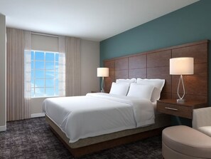 A hotel room with a large bed, two bedside tables with lamps, a wooden headboard, a window with curtains, and a carpeted floor.