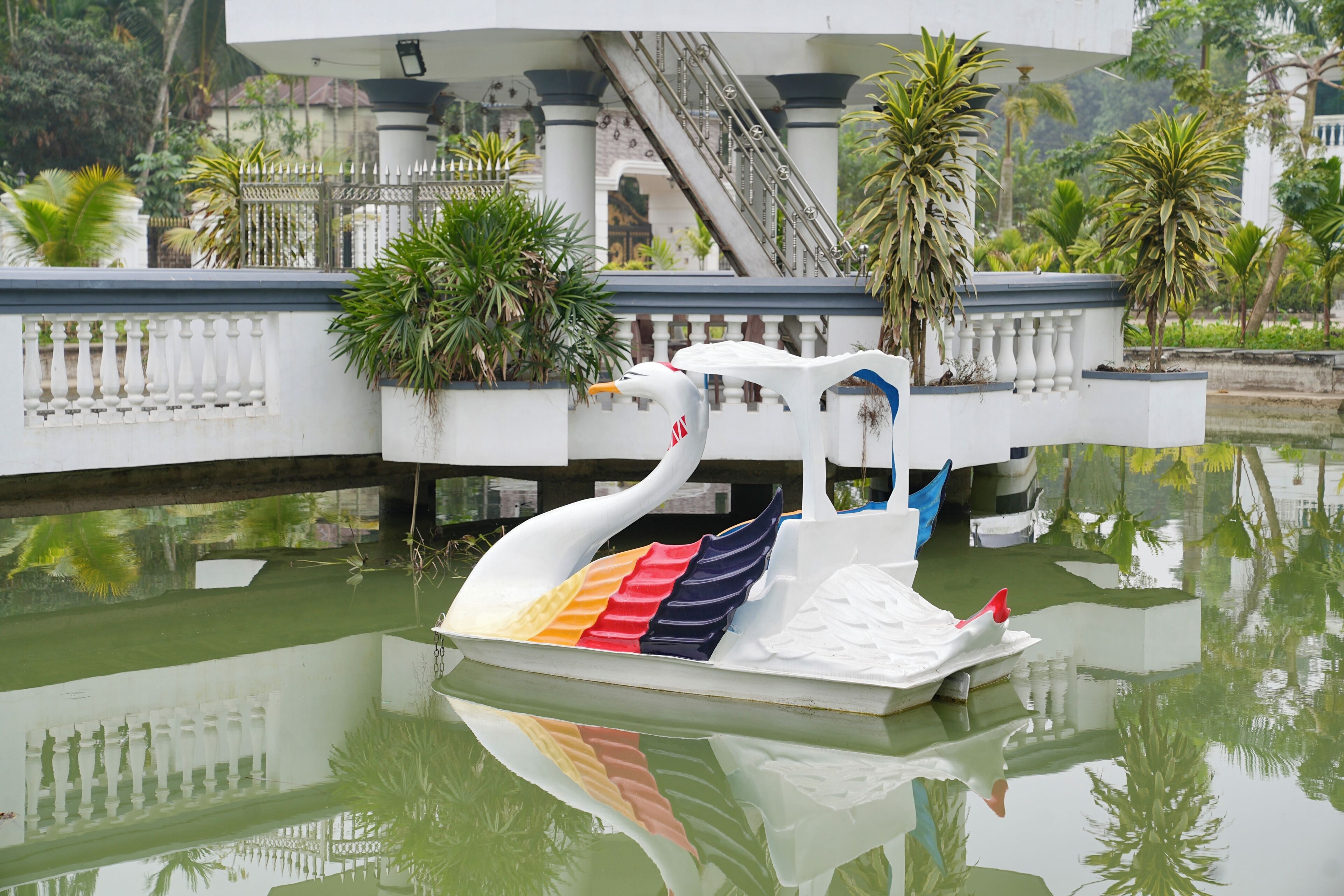 Um barco em forma de cisne em águas calmas, com um edifício branco e palmeiras ao fundo.