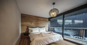 Un dormitorio moderno con cabecera de madera, una cama grande con ropa blanca, una lámpara colgante y un balcón con barandilla de metal.