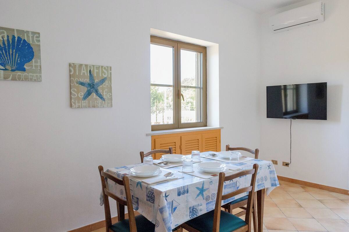 A dining area with a table set for four, a window with shutters, and a mounted TV on the wall.