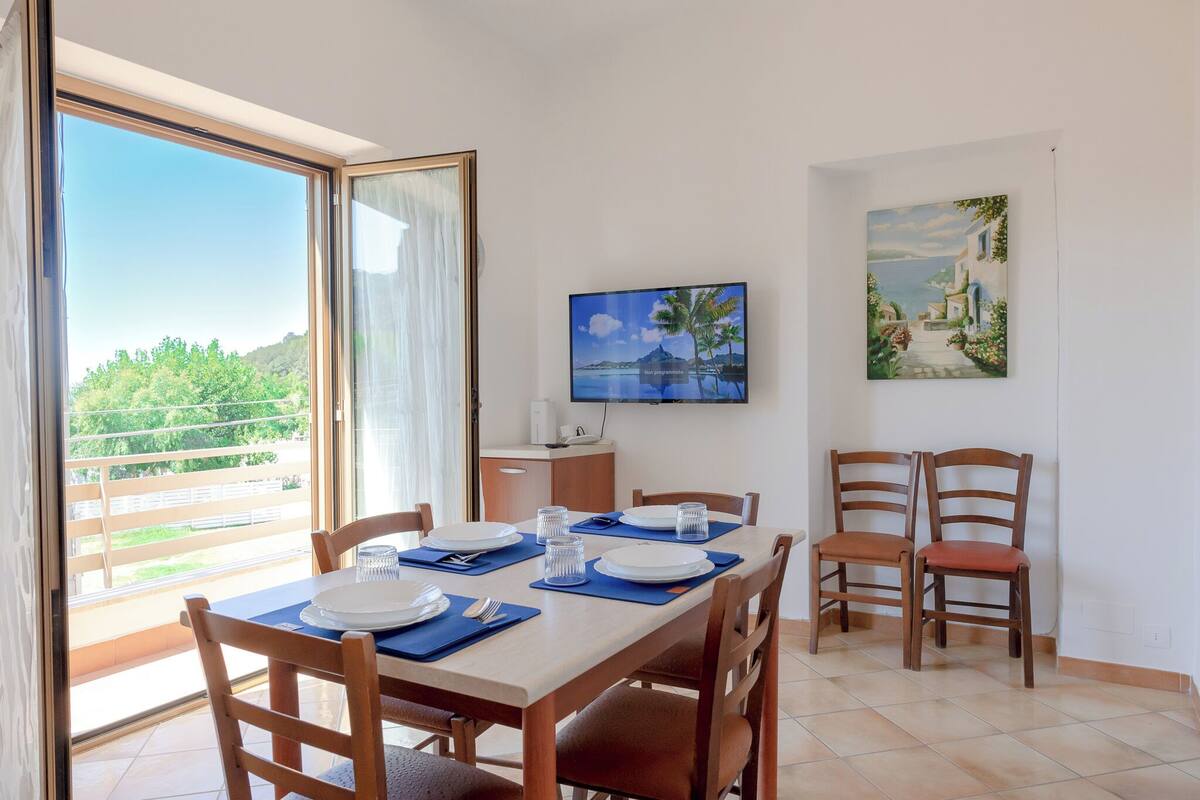A dining area with a table set for two, a TV mounted on the wall, and a painting above the table.