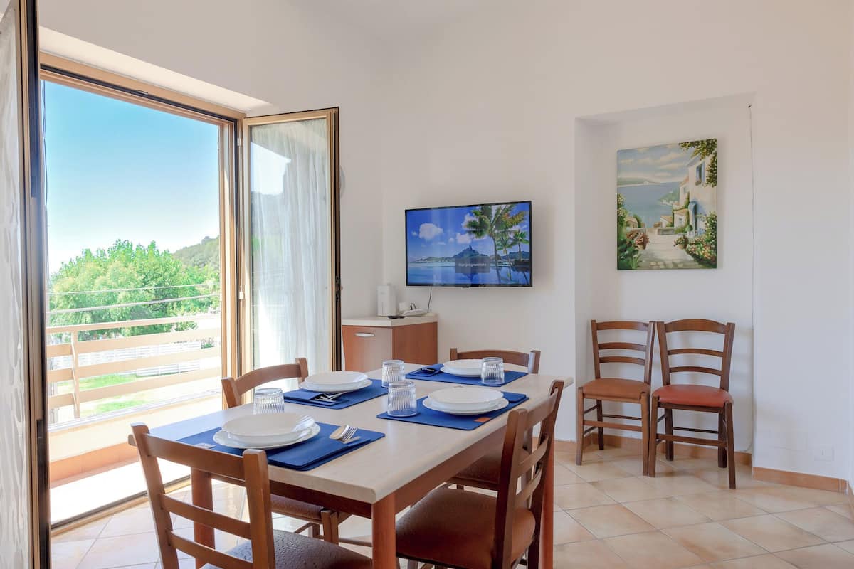 A dining area with a table set for two, a TV mounted on the wall, and a painting above the table.